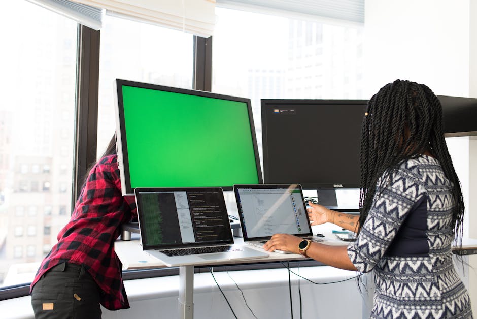 Two women working on laptops and monitors in a bright office setting.